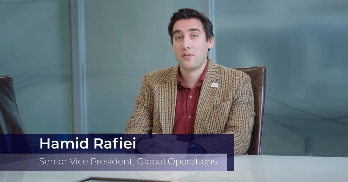 A video still of Hamid Rafiei, Senior Vice President of Global Operations, speaking at a table in a professional office environment.