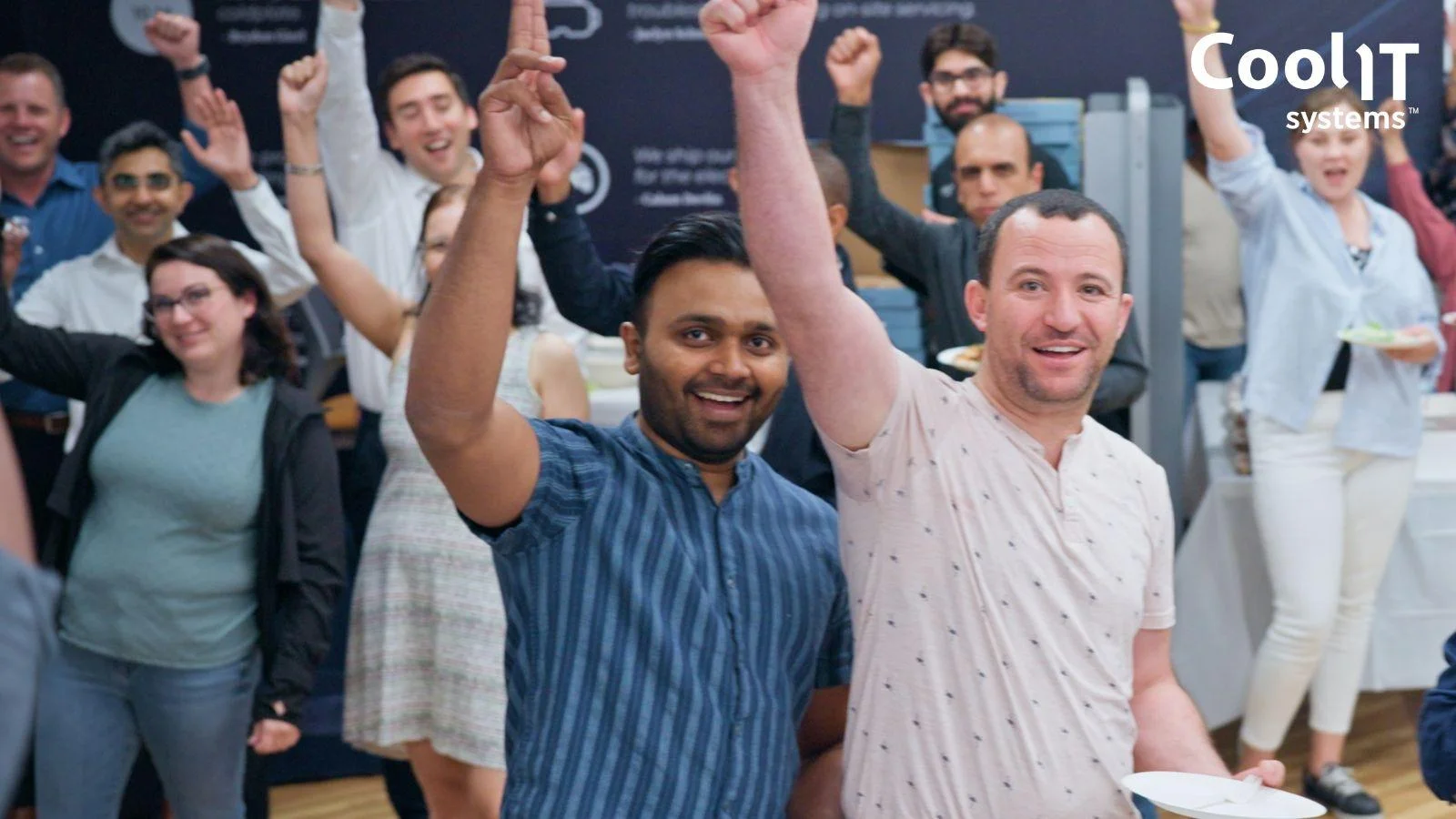 A large group of diverse employees at CoolIT Systems cheering and raising their fists in celebration during an indoor company gathering.