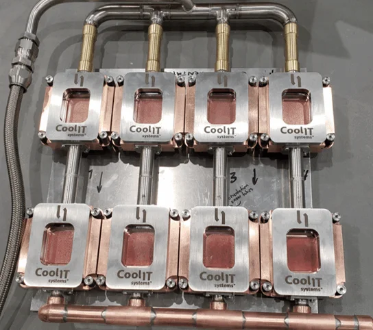 A close-up view of eight silver metal modules with "CoolIT Systems" branding, mounted on a copper base and interconnected by silver and brass piping.