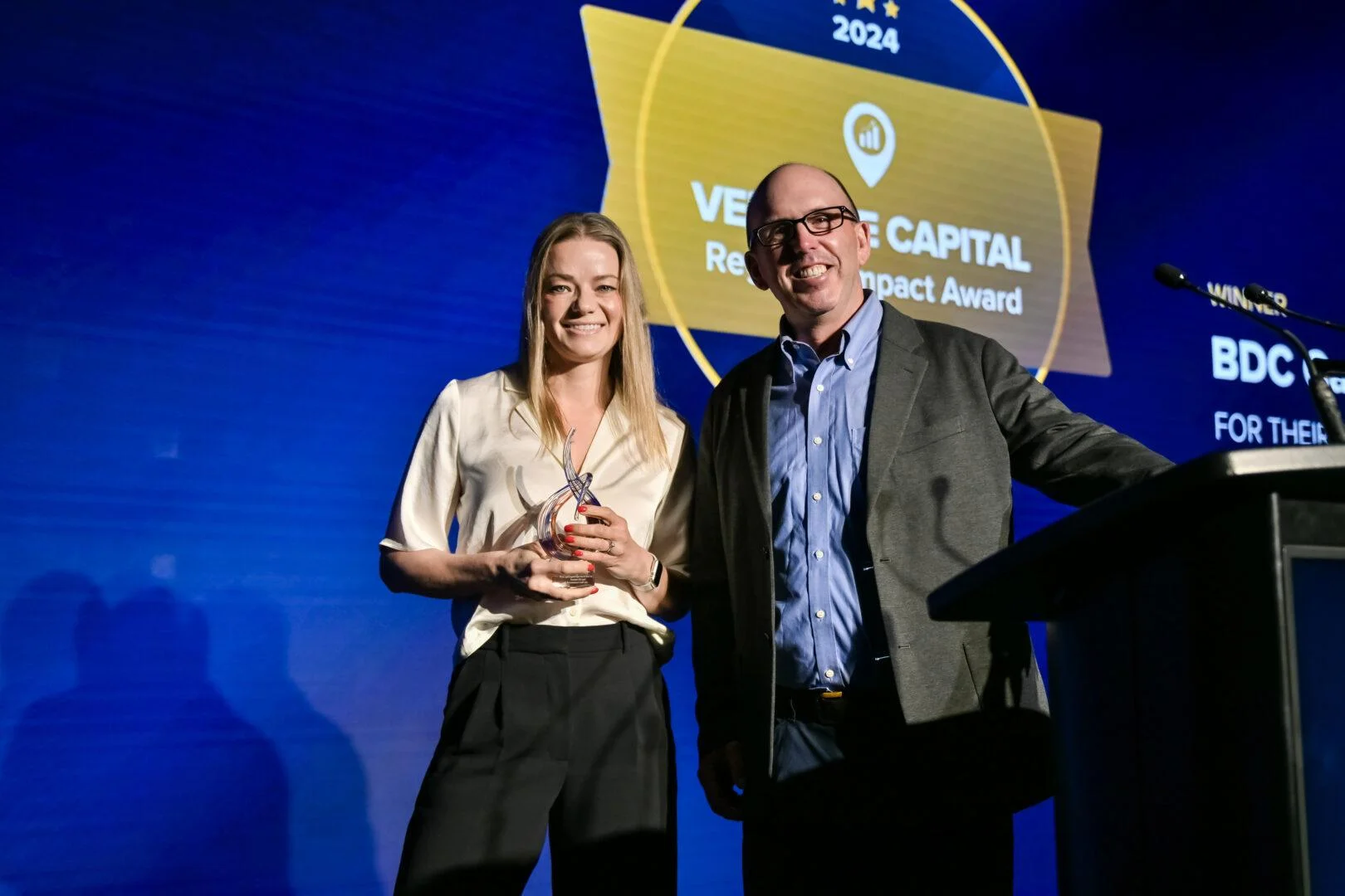 A man and a woman standing together on a stage in front of a blue digital backdrop; the woman is holding a glass trophy, and the backdrop displays the text "VENTURE CAPITAL Regional Impact Award 2024."