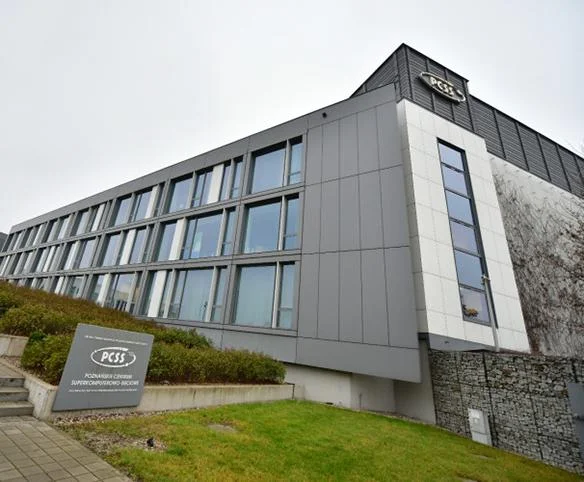 An exterior shot of the Poznań Supercomputing and Networking Center (PCSS) building, showing its modern grey and white tiered architectural design and external signage.