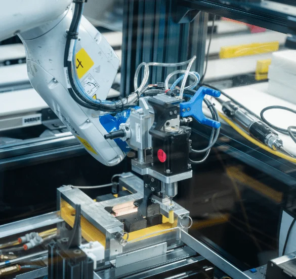 A close-up of a white robotic assembly arm with a specialized black tool attachment working on a yellow hardware component within a high-tech production cell.