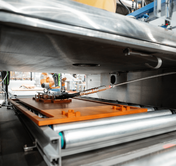 A view from inside a large piece of industrial machinery, showing a hardware component on a wooden platform moving along a metal roller conveyor system.