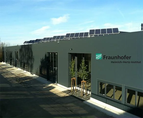 A daytime exterior view of the Fraunhofer Heinrich-Hertz-Institut building, featuring a dark grey facade with solar panels installed across the roofline.