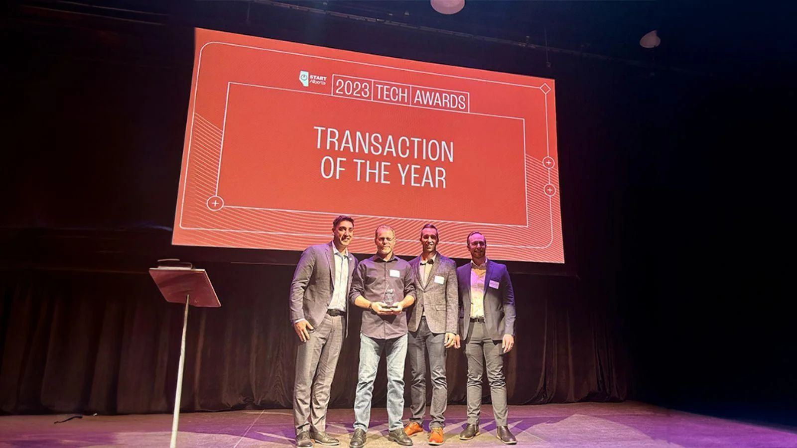 Four men in professional attire posing together on a stage in front of a large red projection screen displaying "2023 TECH AWARDS: TRANSACTION OF THE YEAR."