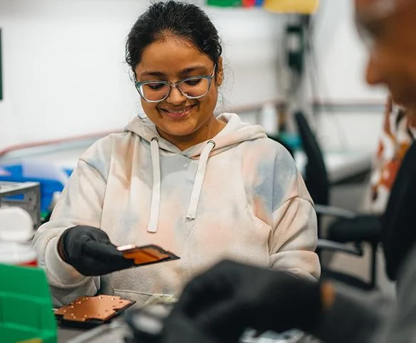 A woman with glasses and black gloves smiles while carefully inspecting a small copper cooling component in a technical laboratory setting.