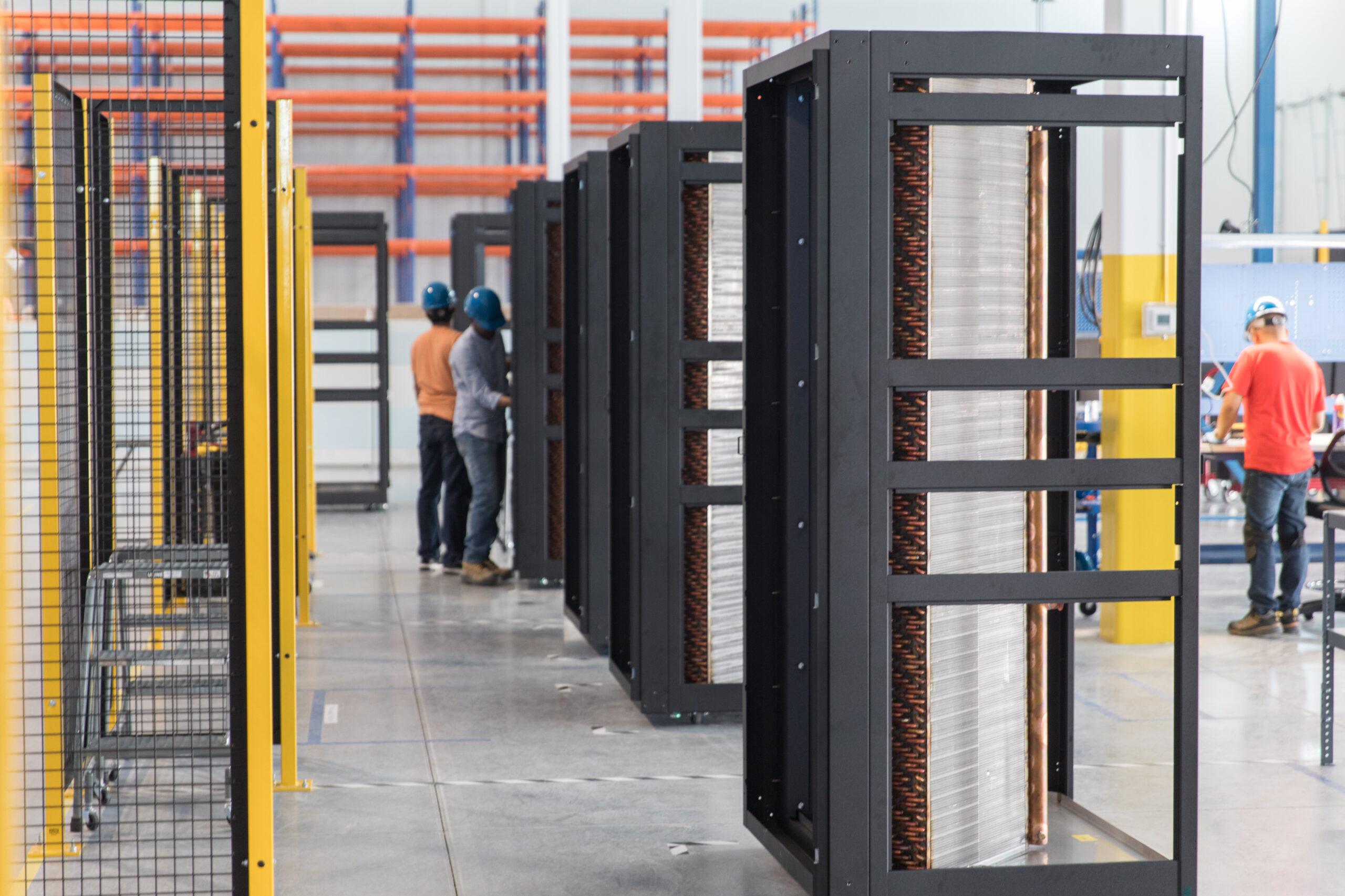 A wide perspective shot inside a factory where several black industrial cabinet frames are lined up for assembly, with technicians in hard hats working in the background.
