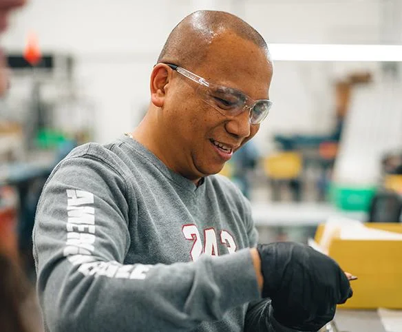 A man wearing safety glasses and a grey long-sleeved shirt smiles while working with his hands on a task in a manufacturing or laboratory environment.