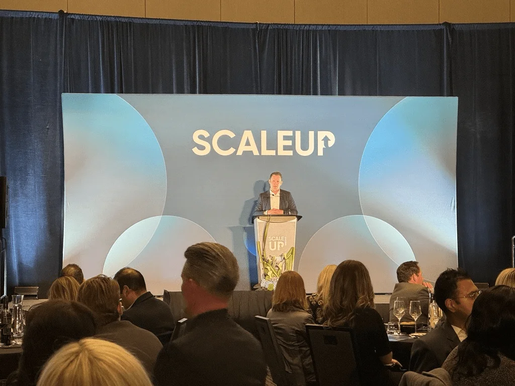 A view of a presenter at a "SCALEUP" event podium, captured from the perspective of the audience sitting at tables in a dimly lit ballroom.