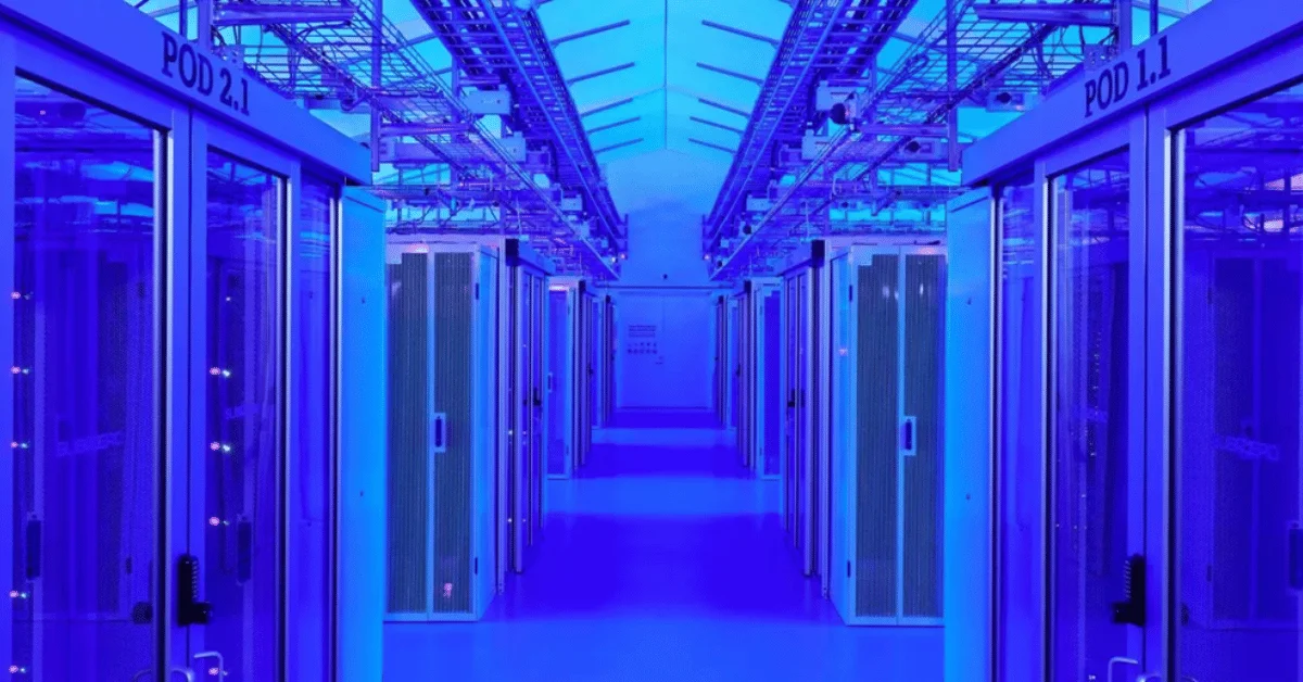 A long perspective shot down a data center aisle illuminated with intense blue light, featuring rows of server containment pods labeled "POD 2.1" and "POD 1.1" with overhead cable management.