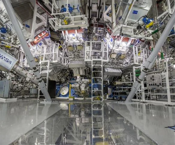 A ground-level view inside a massive, high-tech scientific facility with a complex industrial structure of white beams, platforms, and heavy machinery reflected in a polished floor.