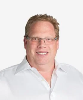 A professional headshot of Ian Graham, smiling and wearing glasses with a white button-down shirt.