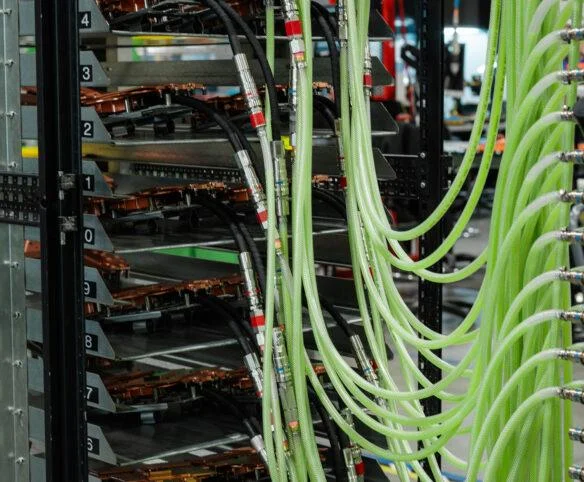 A tall industrial testing rack holding multiple horizontal hardware units, with dozens of translucent green hoses and black cables connected to the front of the assembly.
