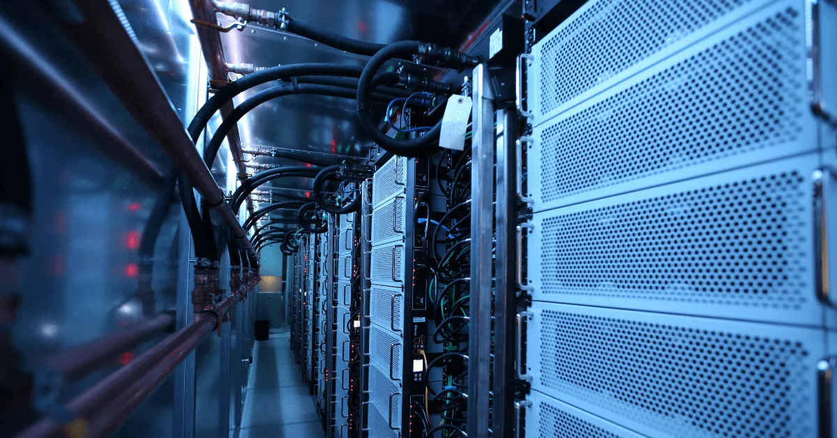 An interior view of a data center showing a dense row of server equipment with white perforated panels and an intricate network of black hoses and copper pipes above.