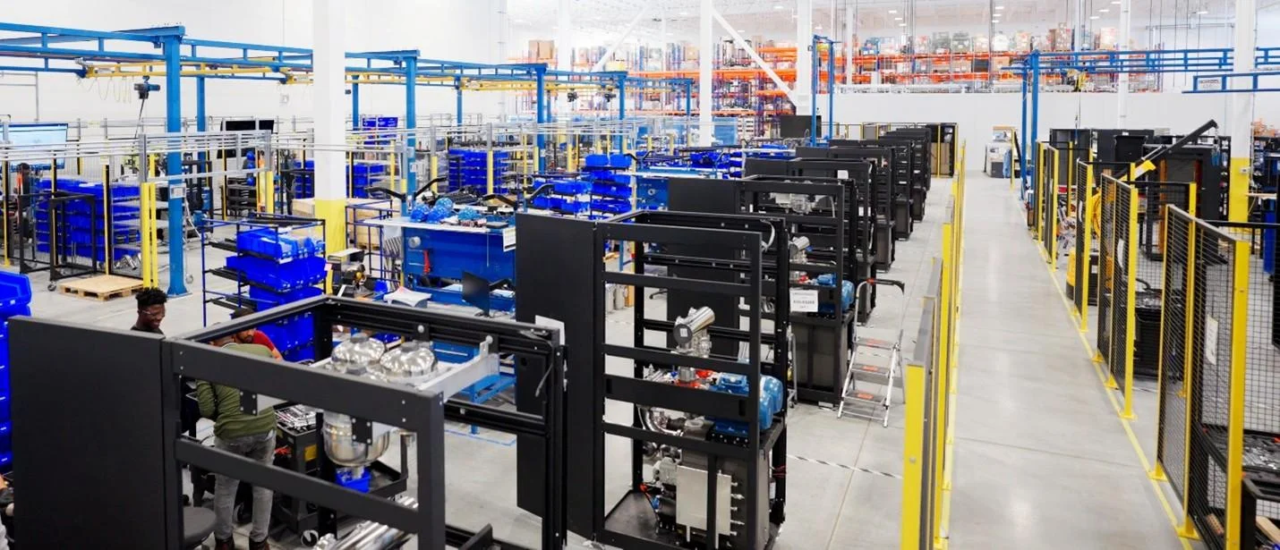 A wide-angle view of a large, brightly lit industrial manufacturing floor with multiple rows of equipment frames, blue storage bins, and workstations in various stages of assembly.
