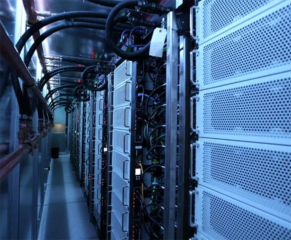 An interior view of a data center aisle featuring rows of server equipment with white perforated panels and a complex network of black cooling hoses and copper pipes overhead.