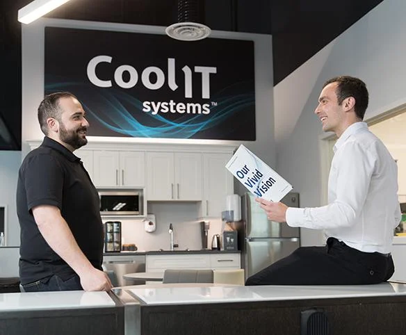 Two men in professional attire are having a friendly conversation in a modern office kitchen area; one man is sitting on a counter holding a brochure titled "Our Vivid Vision" while the CoolIT Systems logo is visible on the wall behind them.