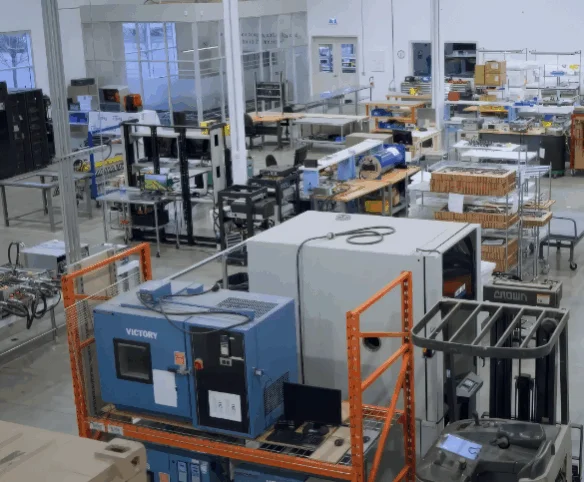 A high-angle view of a bright industrial laboratory or testing facility filled with various workstations, electronic testing equipment, and large blue machinery.