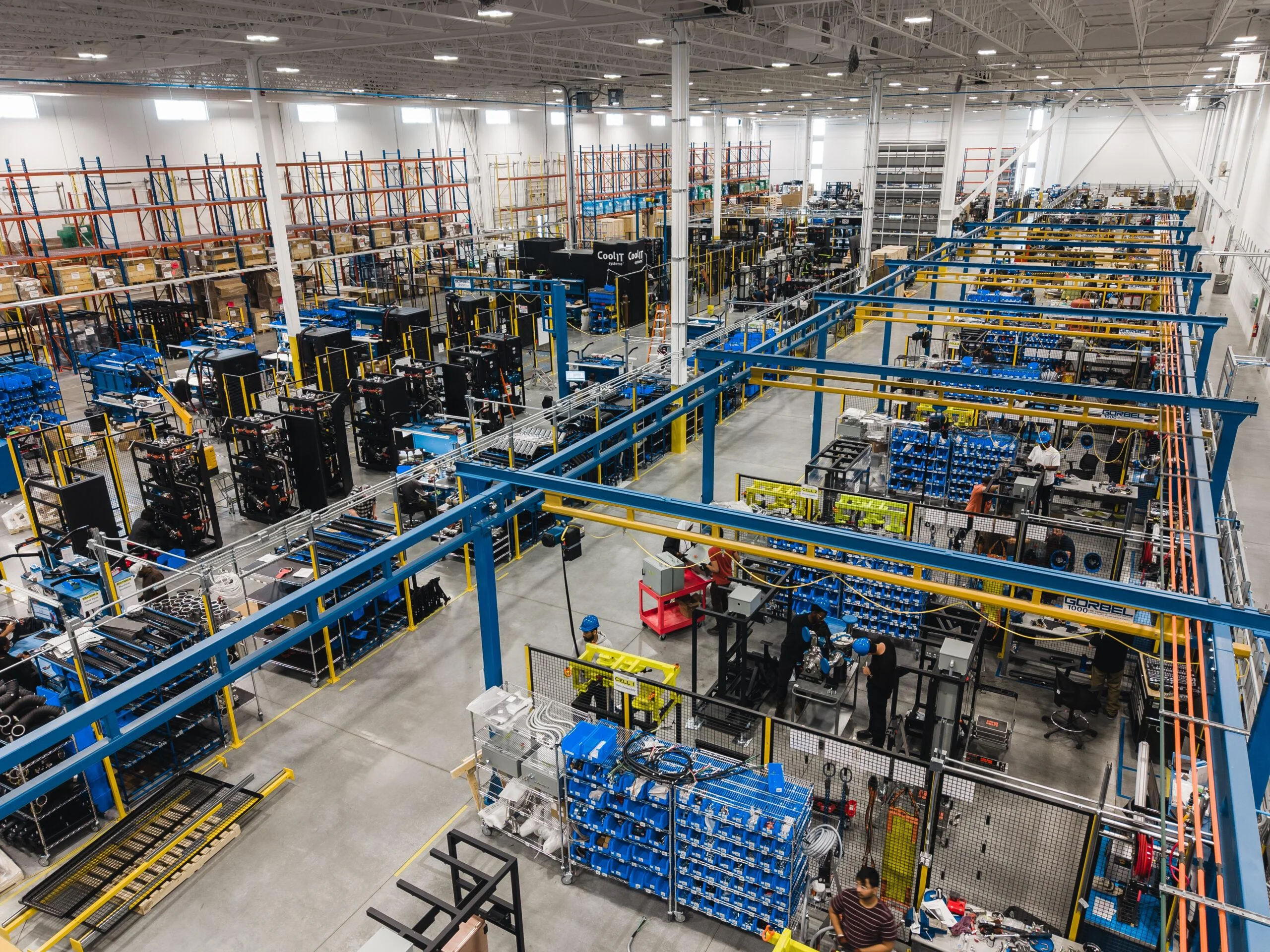 An overhead wide-angle view of a vast and busy manufacturing floor, featuring organized production cells, blue overhead frames, and technicians assembling large-scale industrial hardware.