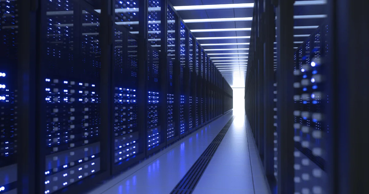 A long perspective view down a dark data center corridor lined with server racks glowing with blue LED lights, leading toward a bright white light at the end.