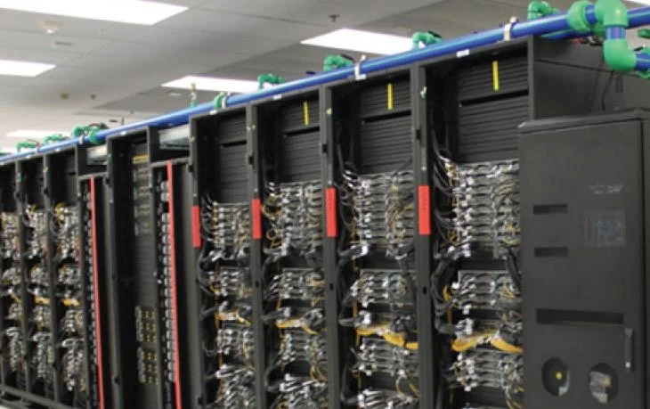 Liquid-cooled data centre racks with overhead coolant piping and high-density server hardware.