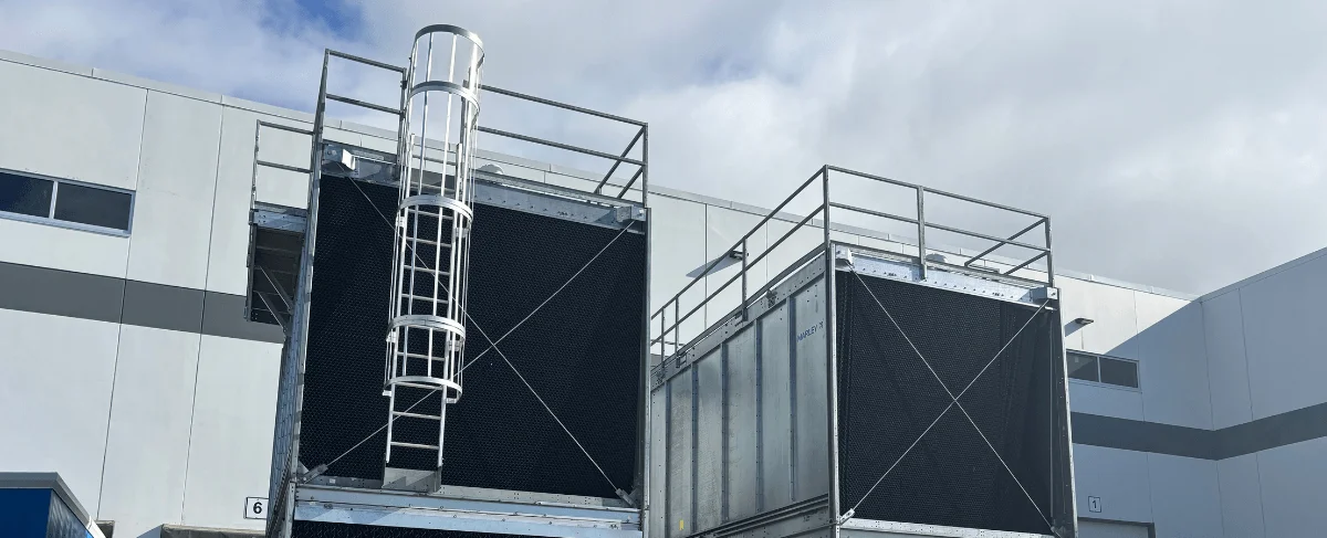 Two large industrial cooling units with protective railings and a ladder are positioned outside a building.