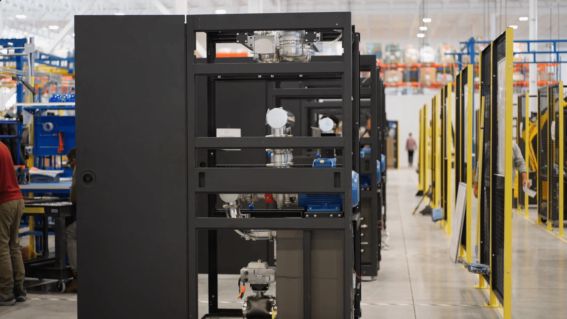 A view down a central aisle of a manufacturing facility, showing rows of large black equipment frames and yellow safety railings with technicians working at workstations in the background.