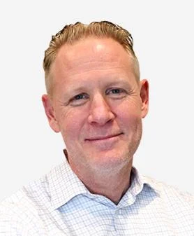 A professional headshot of Adam Schader, smiling and wearing a white checkered button-down shirt.