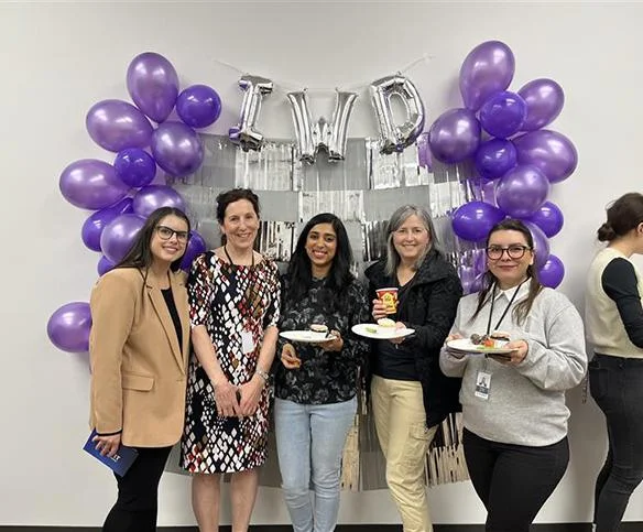 Five women stand together smiling in front of a celebratory wall decorated with purple balloons and silver "IWD" (International Women's Day) letter balloons while holding small plates of food.