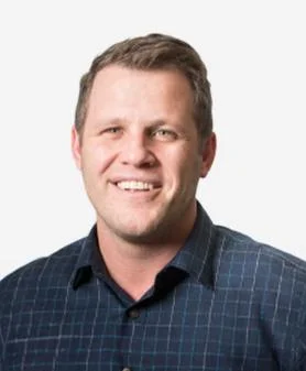 A professional headshot of Patrick McGinn, smiling and wearing a dark checkered button-down shirt.