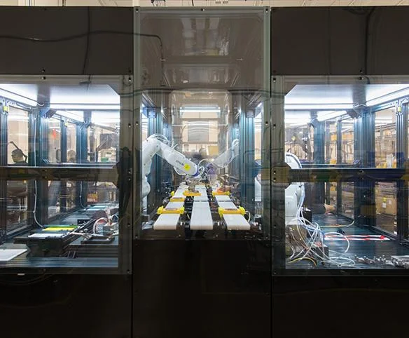 A view through a glass safety enclosure of an automated production line where a white robotic arm is performing assembly tasks on hardware components.