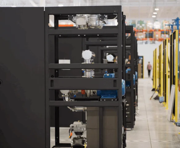 A view down a central aisle of a manufacturing facility, showing rows of large black equipment frames and yellow safety railings with technicians working at workstations in the background.