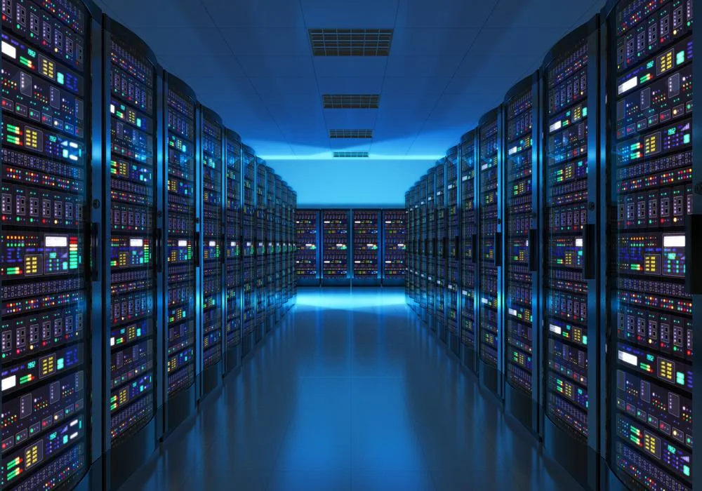 A long, symmetrical view of a dark data center aisle with rows of server racks illuminated by bright blue floor and ceiling lights, with visible status indicator LEDs on the equipment.