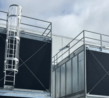 Two large industrial cooling units with protective railings and a ladder are positioned outside a building.