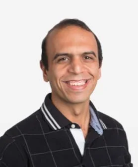 A professional headshot of Kamal Mostafavi, smiling and wearing a black polo shirt with white stripes on the collar.