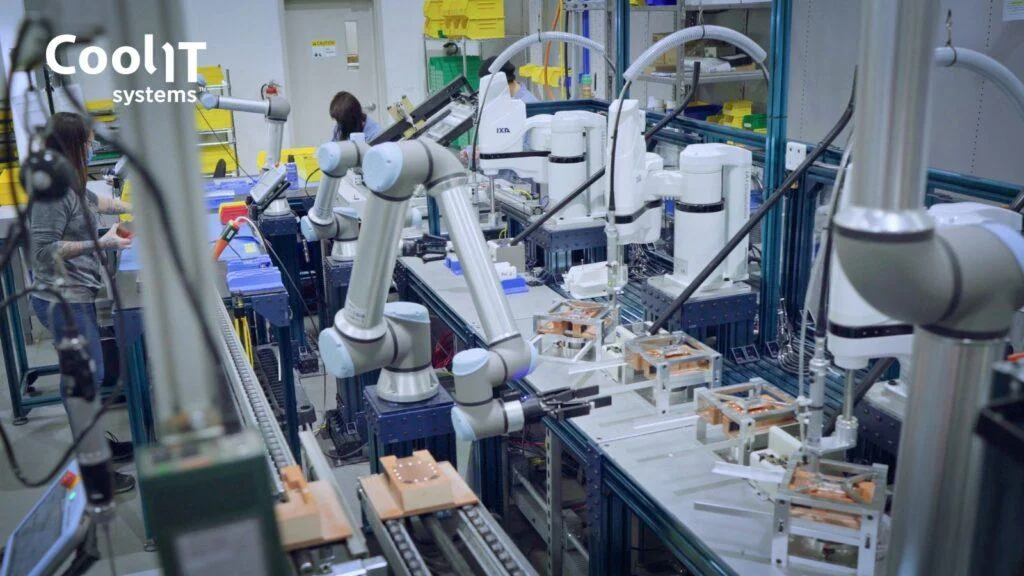 A wide shot of an automated production line where several white robotic arms are positioned over a conveyor system to assemble hardware components, with a technician visible on the left.